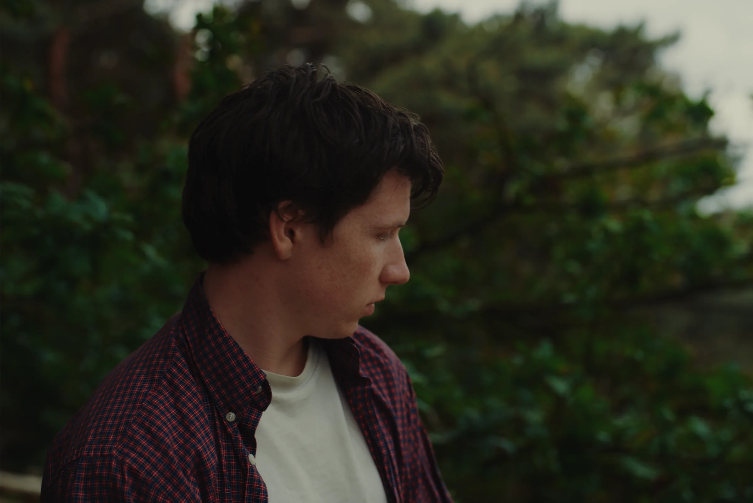 A young man stands outdoors among trees.