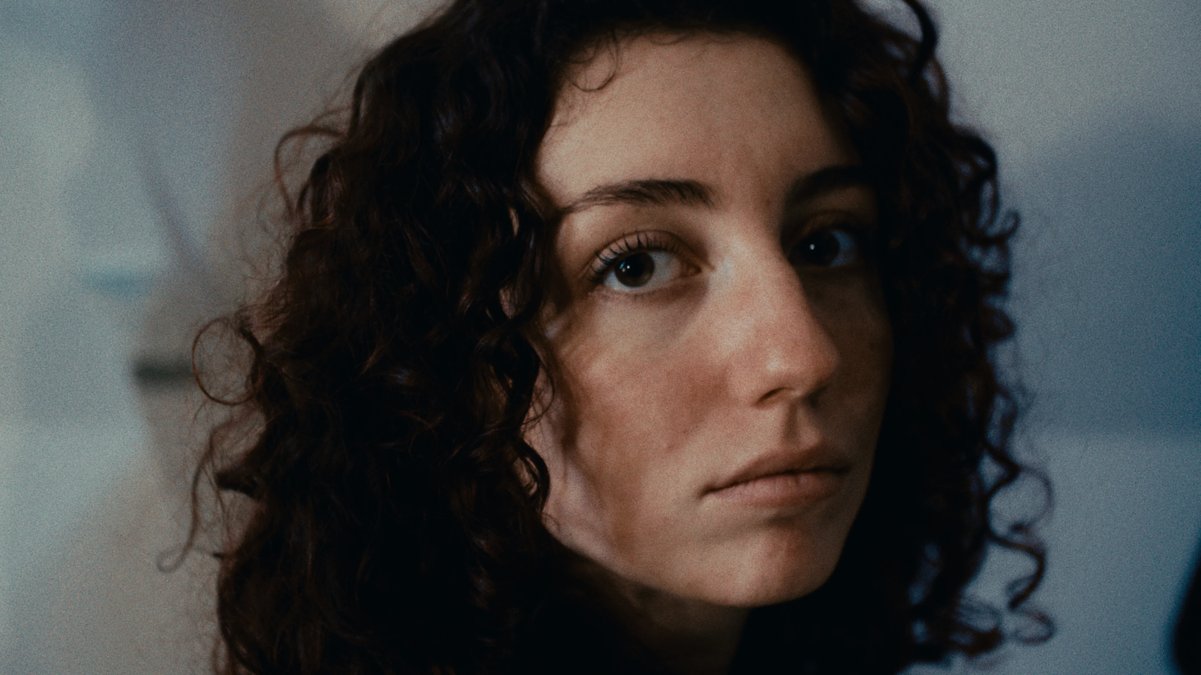 A woman with dark curls looks into the camera against a cool-toned background.