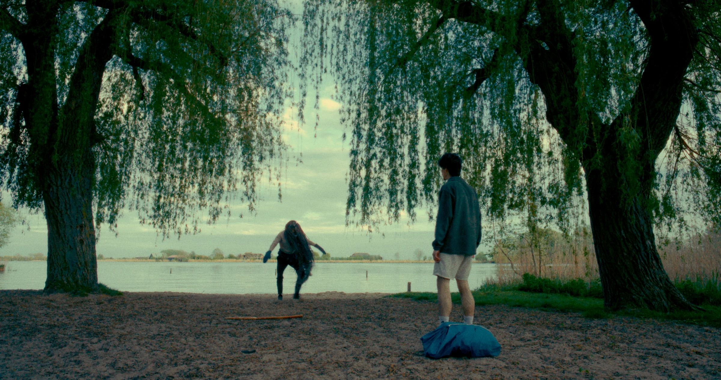 Two figures stand beneath willow trees beside the water.