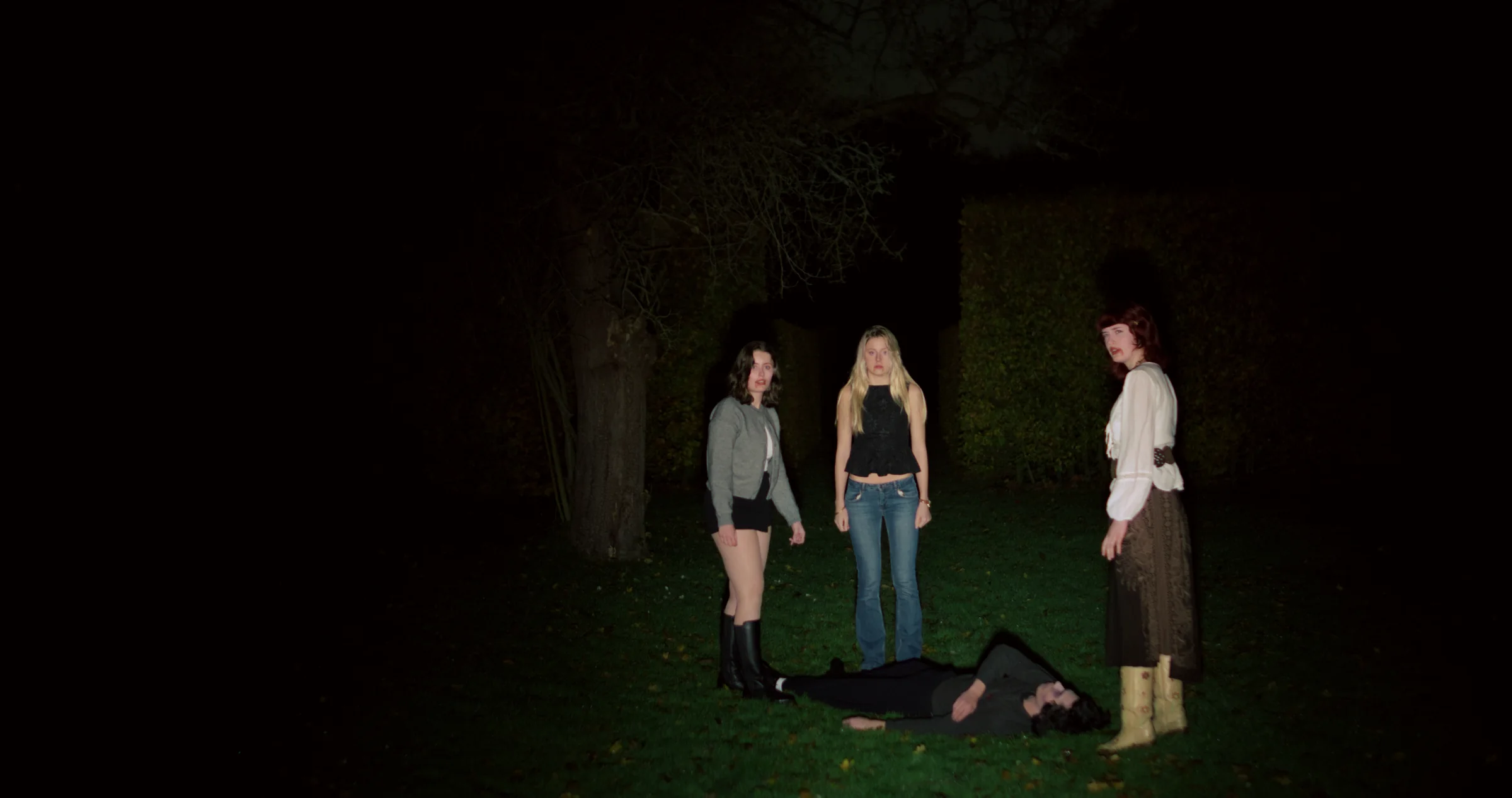 Three figures stand together in a dark wooded setting.