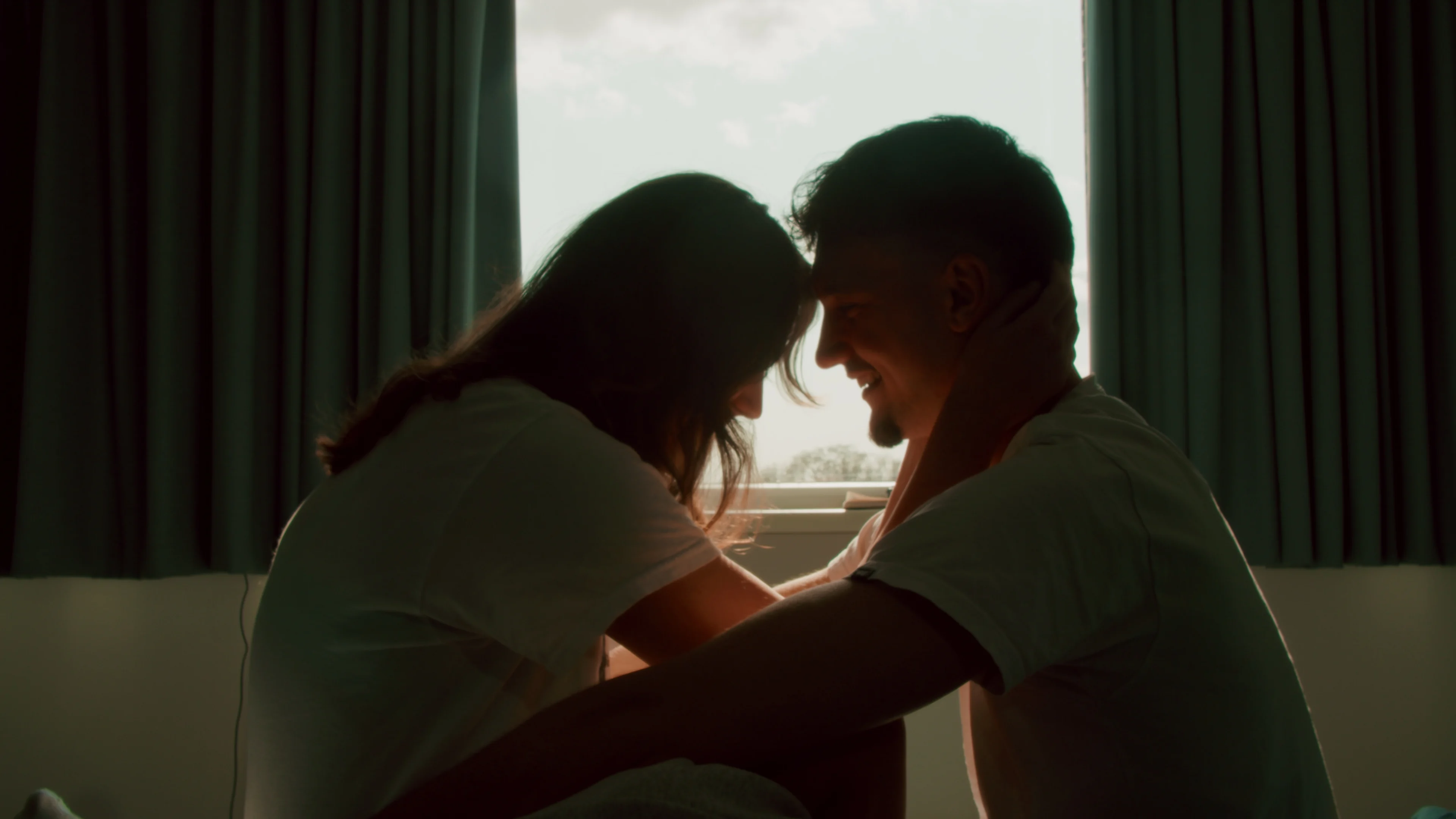 A woman and a man sit close together beside a bright window.